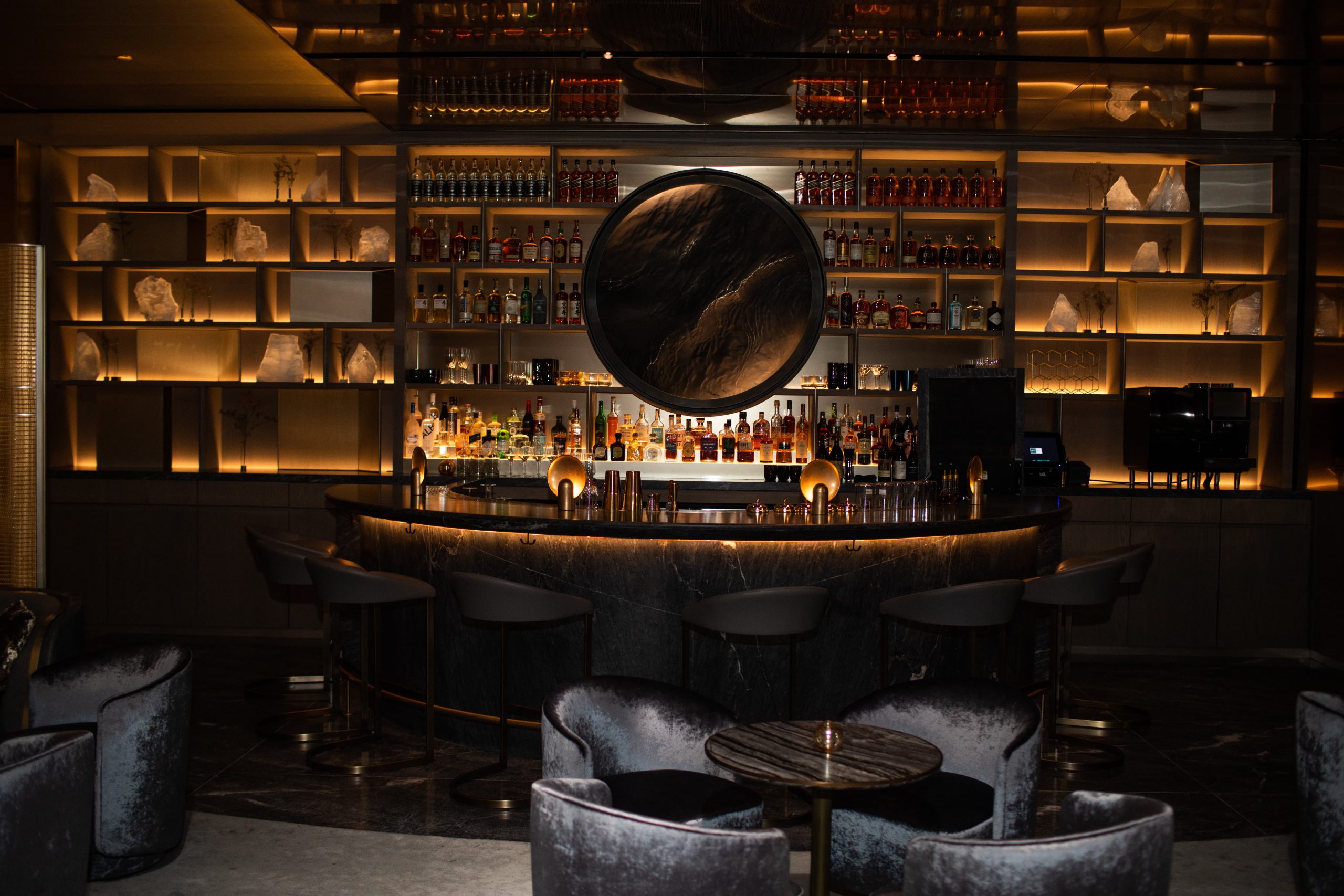 A dimly lit, upscale bar featuring a circular centerpiece mirror, surrounded by shelves stocked with bottles. The bar has high-backed stools and plush chairs in the foreground, creating a cozy, inviting atmosphere.