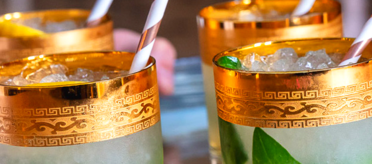 Four glasses with intricate gold rims filled with iced drinks, each with a striped straw and garnished with mint leaves.