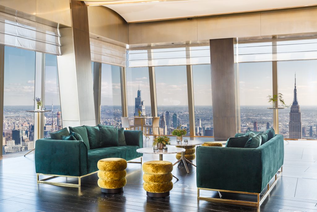 A modern lounge with emerald green sofas and gold velvet stools is set against floor-to-ceiling windows offering a panoramic city skyline view, including a prominent tower. Light wood flooring and sleek decor enhance the elegant atmosphere.