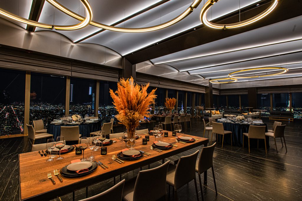 A luxurious, modern dining room with large windows overlooking a night cityscape. The room features elegant circular ceiling lights, wooden tables set with plates and glasses, and tall vases with orange floral arrangements as centerpieces.