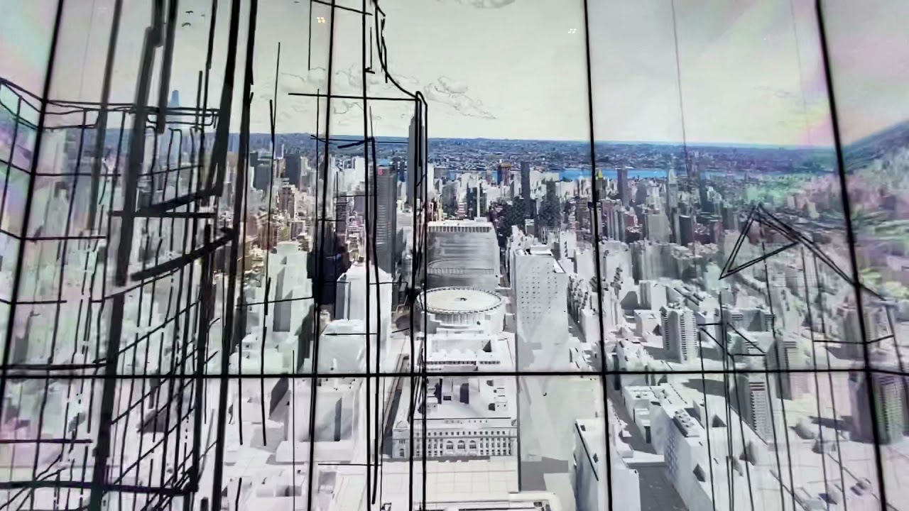 A digital display shows a stylized aerial view of a cityscape with skyscrapers and urban architecture. The image includes black line drawings overlaid on a realistic 3D rendering of the city.