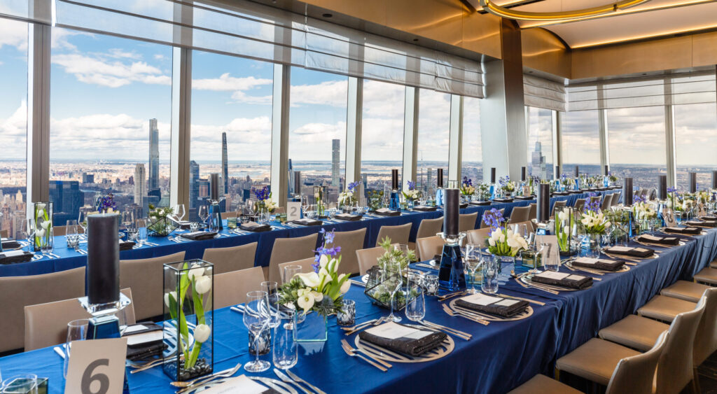 Elegant banquet setup in a high-rise venue with panoramic city views. Long tables are adorned with navy blue tablecloths, white and purple floral centerpieces, black candles, and neatly arranged cutlery. Large windows reveal a skyscraper skyline.