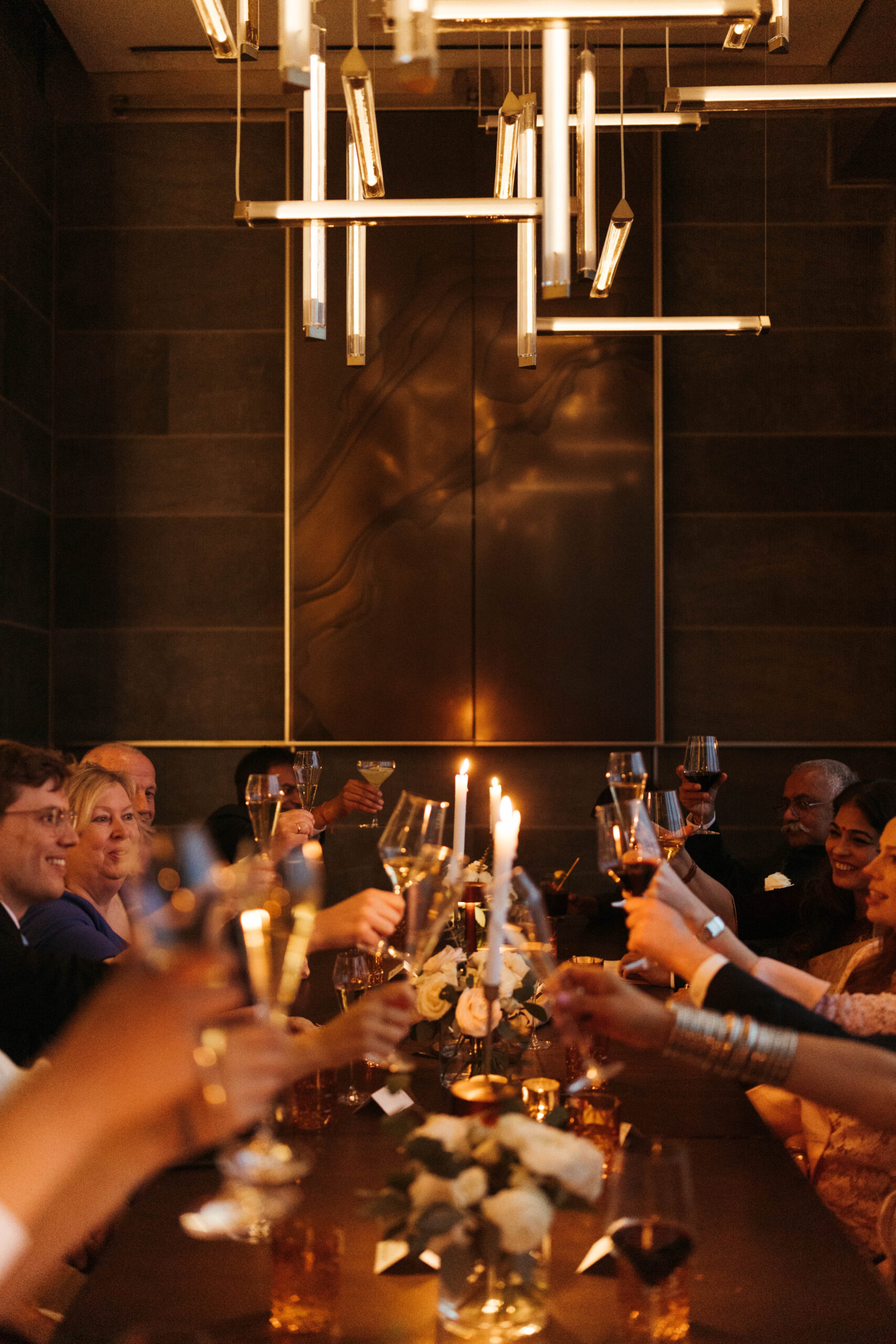 A group of people sitting around a dining table raise their glasses in a toast. The room is dimly lit, with candles and elegant hanging lights. The table is decorated with small flower arrangements.