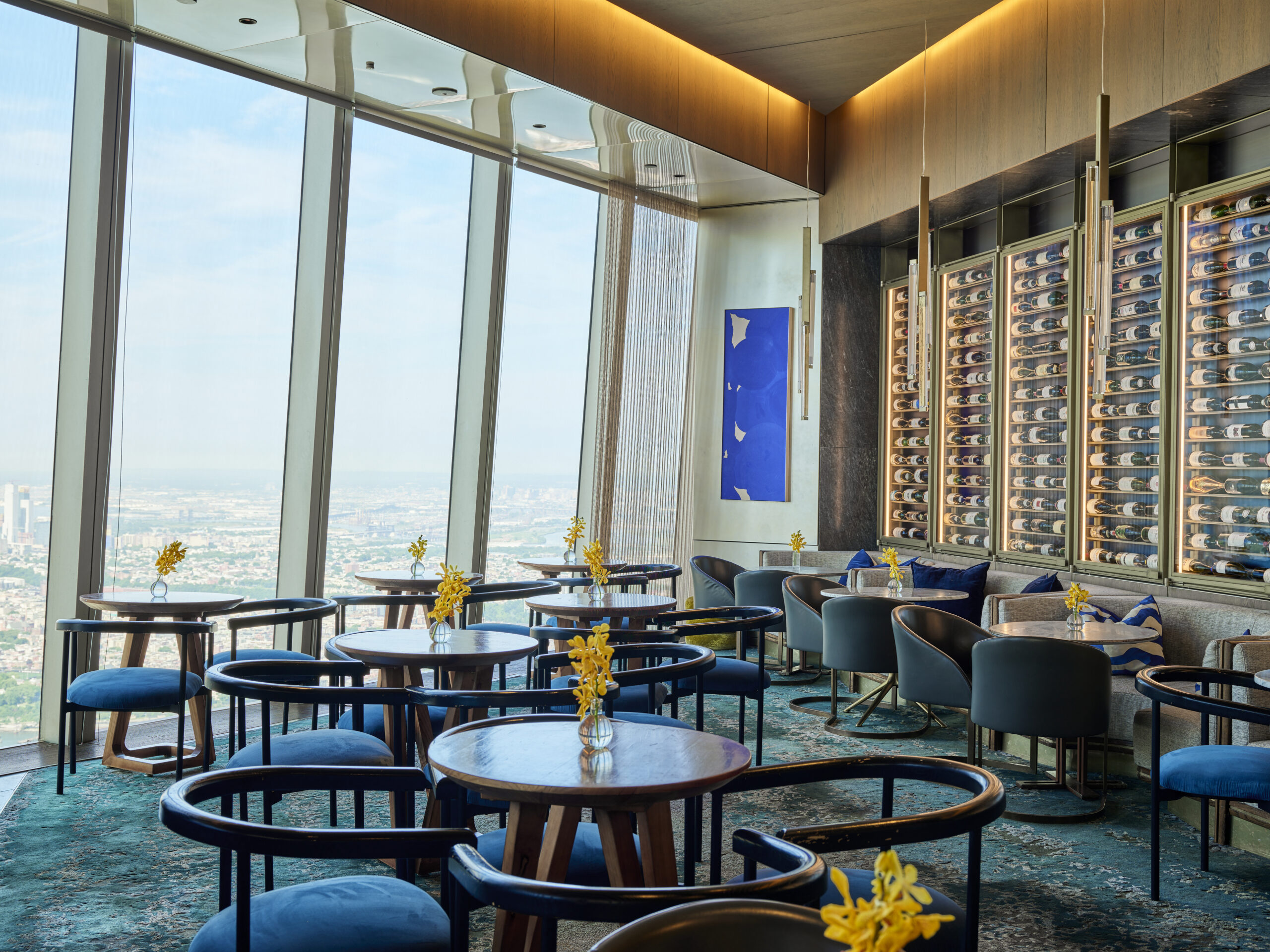 A modern, stylish restaurant with floor-to-ceiling windows offering a panoramic city view. It features round tables with chairs, each table adorned with a small vase of yellow flowers. The interior has contemporary artwork and a wall of wine racks.