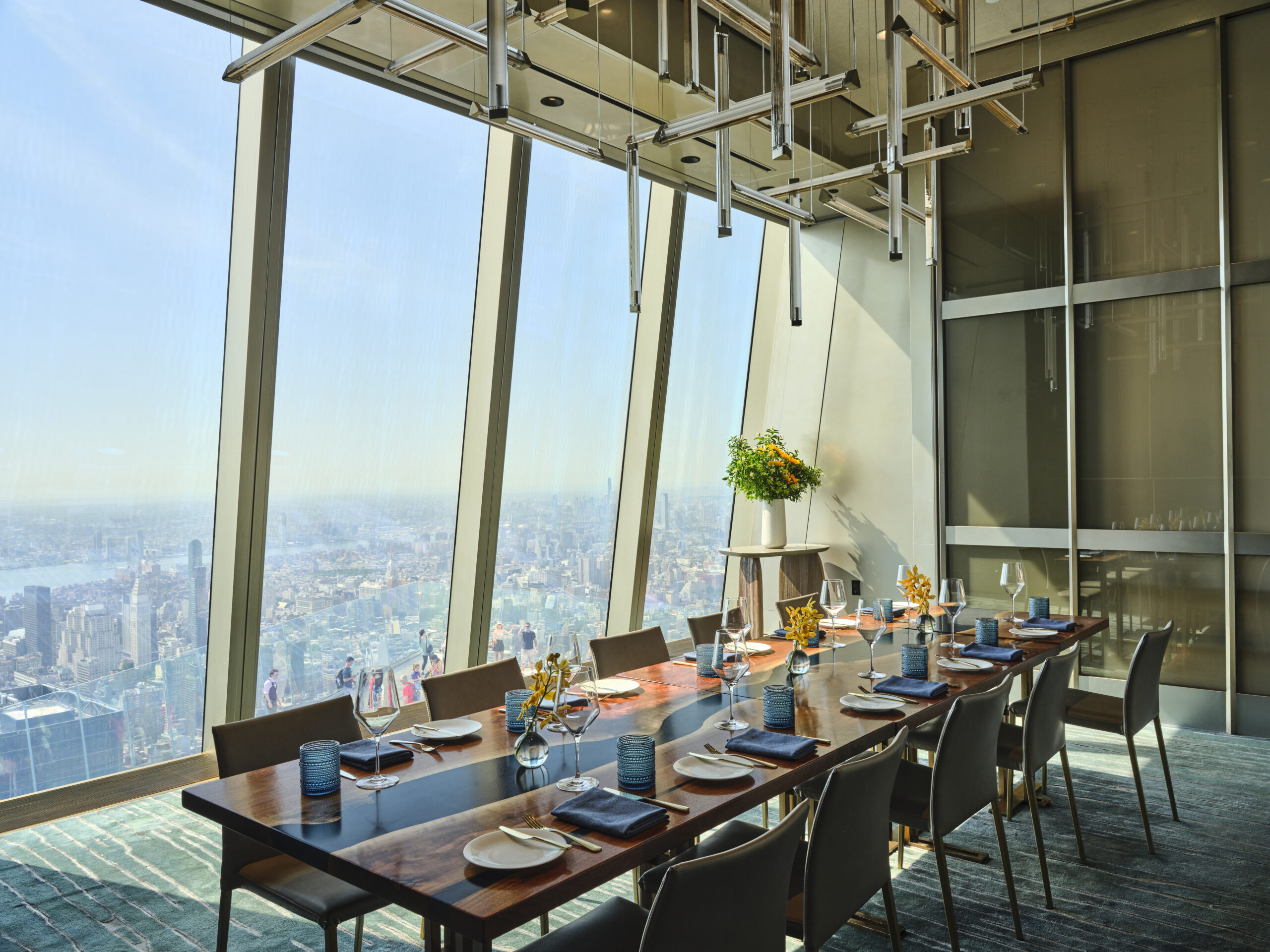 A modern dining room with a long table set for a meal, featuring blue napkins and yellow floral centerpieces. Large floor-to-ceiling windows offer a panoramic view of a cityscape under a clear blue sky.
