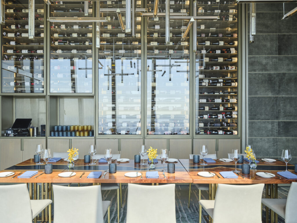 A modern restaurant with a long wooden table set for dining. The table has neatly arranged plates, glasses, and yellow floral centerpieces. Behind, a wall showcases a vast collection of wine bottles.