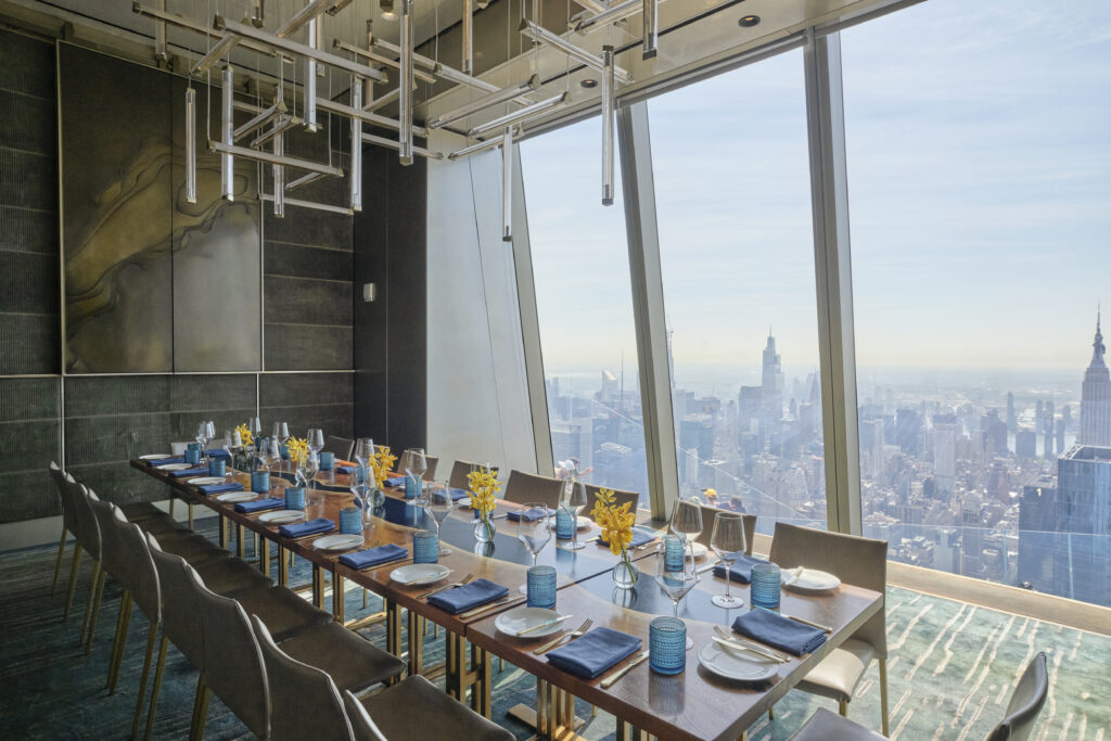 A luxurious dining room with a long table set for a meal, overlooking a cityscape through large floor-to-ceiling windows. Modern lighting hangs from the ceiling, and the table is adorned with blue accents and yellow floral arrangements.