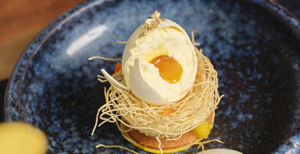 A gourmet dessert resembling a poached egg sits atop a nest of spun sugar on a blue ceramic plate. The dessert has an outer shell, creamy filling, and a yolk-like center, accented with a small piece of gold leaf.