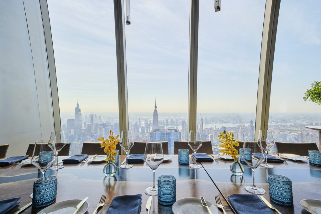 Elegant dining setup by a large window with a panoramic view of a city skyline, including skyscrapers and a clear sky. The table features yellow flower centerpieces, blue glassware, and neatly arranged silverware.