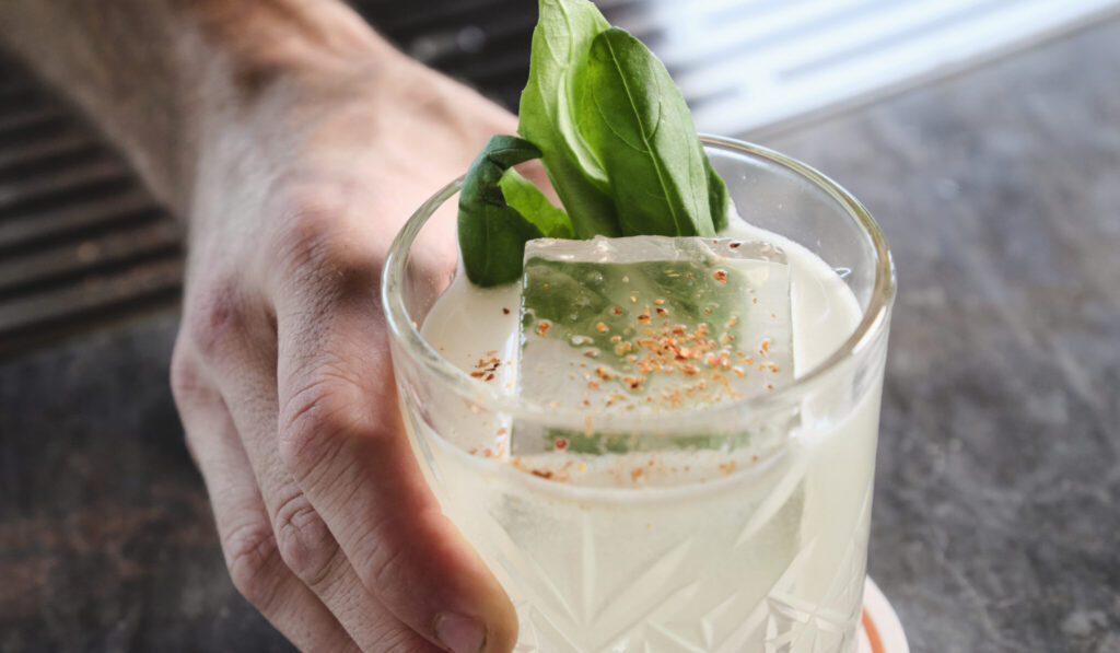 A hand holds a glass filled with a light-colored cocktail. The drink is garnished with a large ice cube, green basil leaves, and a sprinkle of spices on top. The background features a textured surface and part of a metal grate.