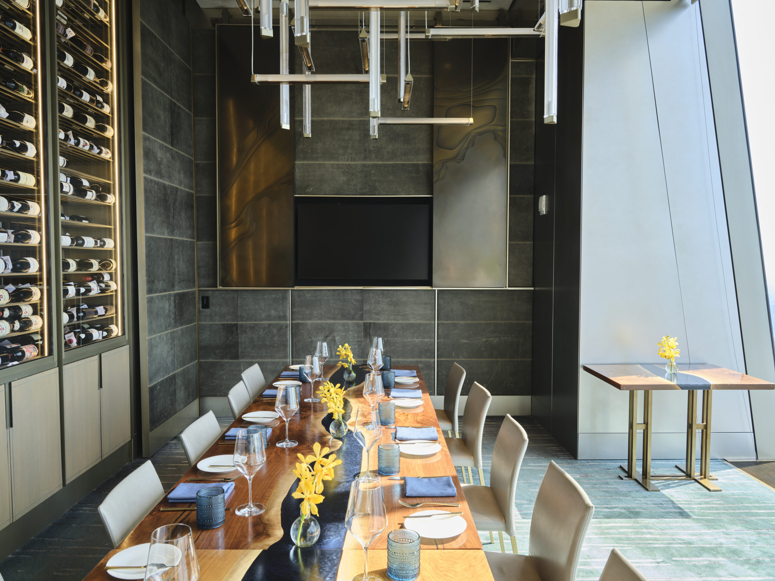 A modern dining room with a long wooden table set for a meal, featuring yellow flowers in vases. The room has a floor-to-ceiling wine rack, a flat-screen TV on the far wall, and large angled windows allowing natural light to filter in.