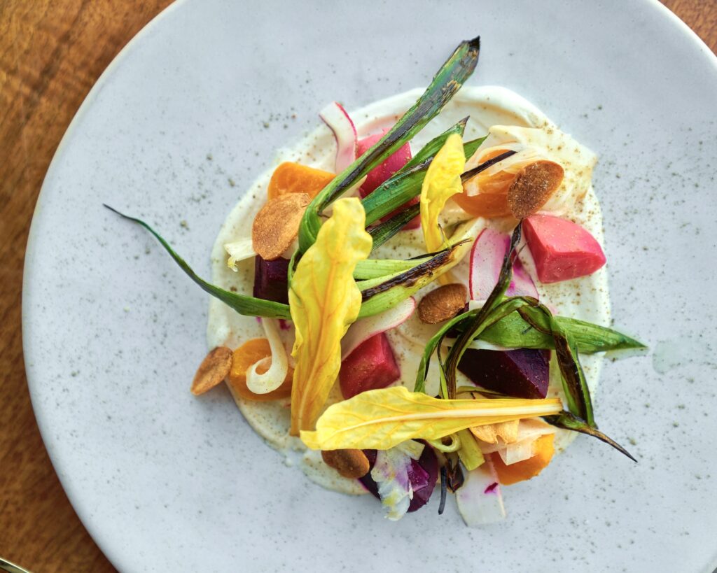 A colorful dish elegantly arranged on a white plate, featuring a variety of fresh vegetables like beets, spring onions, and radishes, garnished with vibrant yellow flower petals and thinly sliced pieces, served on a creamy white sauce.