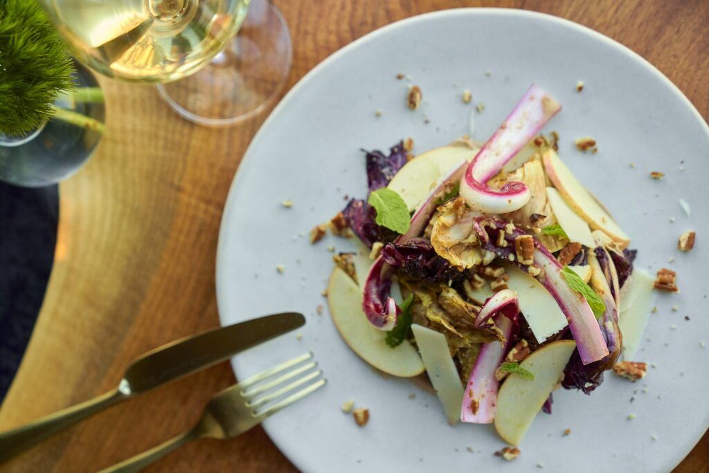 A gourmet salad with endive, apple slices, nuts, and cheese on a white plate. A fork and knife are placed beside the plate on a wooden table, with a glass of white wine nearby.