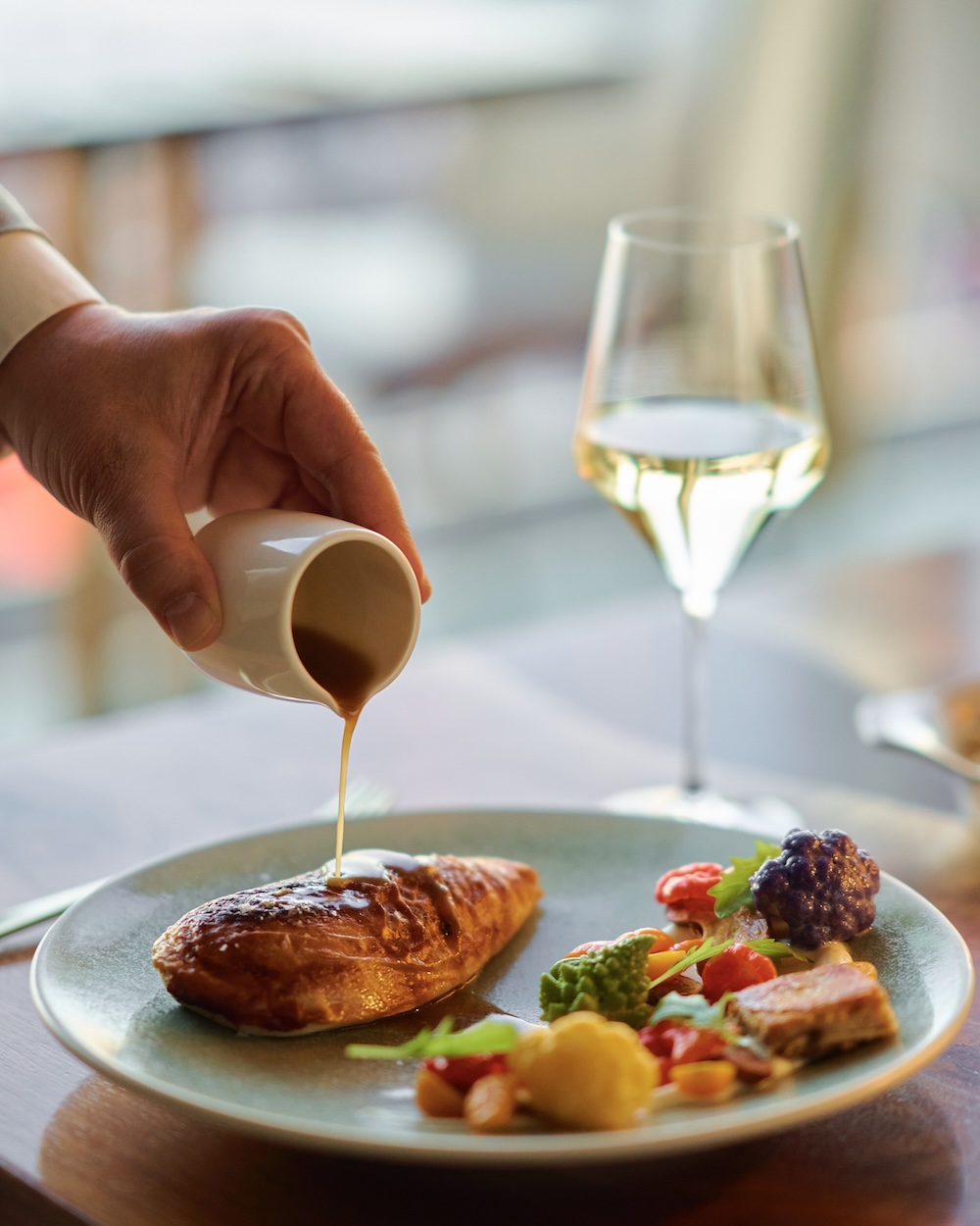 A hand pours sauce over a piece of meat on a plate with colorful vegetables. A glass of white wine is in the background on a table. The setting is a warmly lit dining area, evoking a sophisticated and appetizing atmosphere.