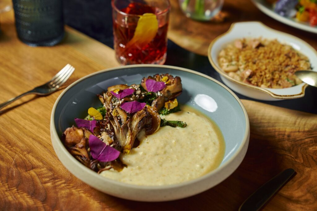 A bowl of creamy polenta topped with roasted mushrooms and purple edible flowers. A reddish drink with an orange slice is in the background, along with a dish of beans and grains. The setting is on a wooden table with cutlery.