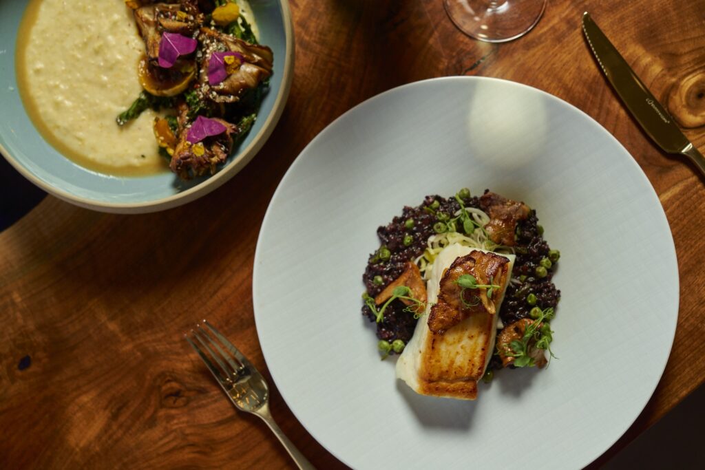 A wooden table with two plates of gourmet food: one with a dish featuring seared fish on grains with herbs, and another with a creamy dish topped with artichokes and colorful garnishes. Nearby are a fork and a wine glass.