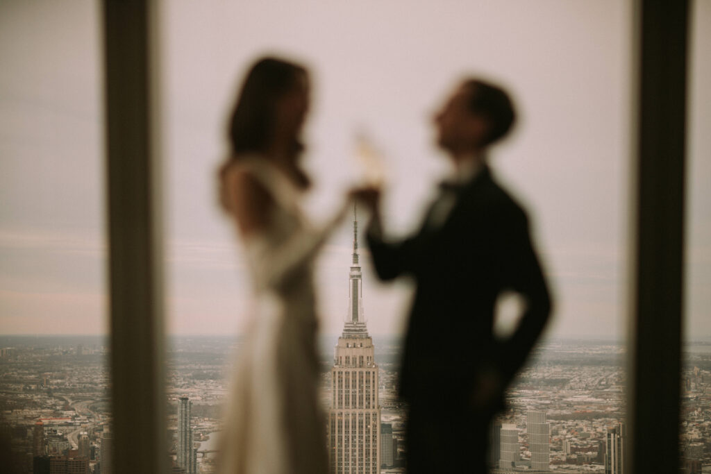 A silhouetted couple toasts in front of an expansive window with the Empire State Building visible in the background. The scene is romantically blurred, highlighting the iconic New York City skyline.