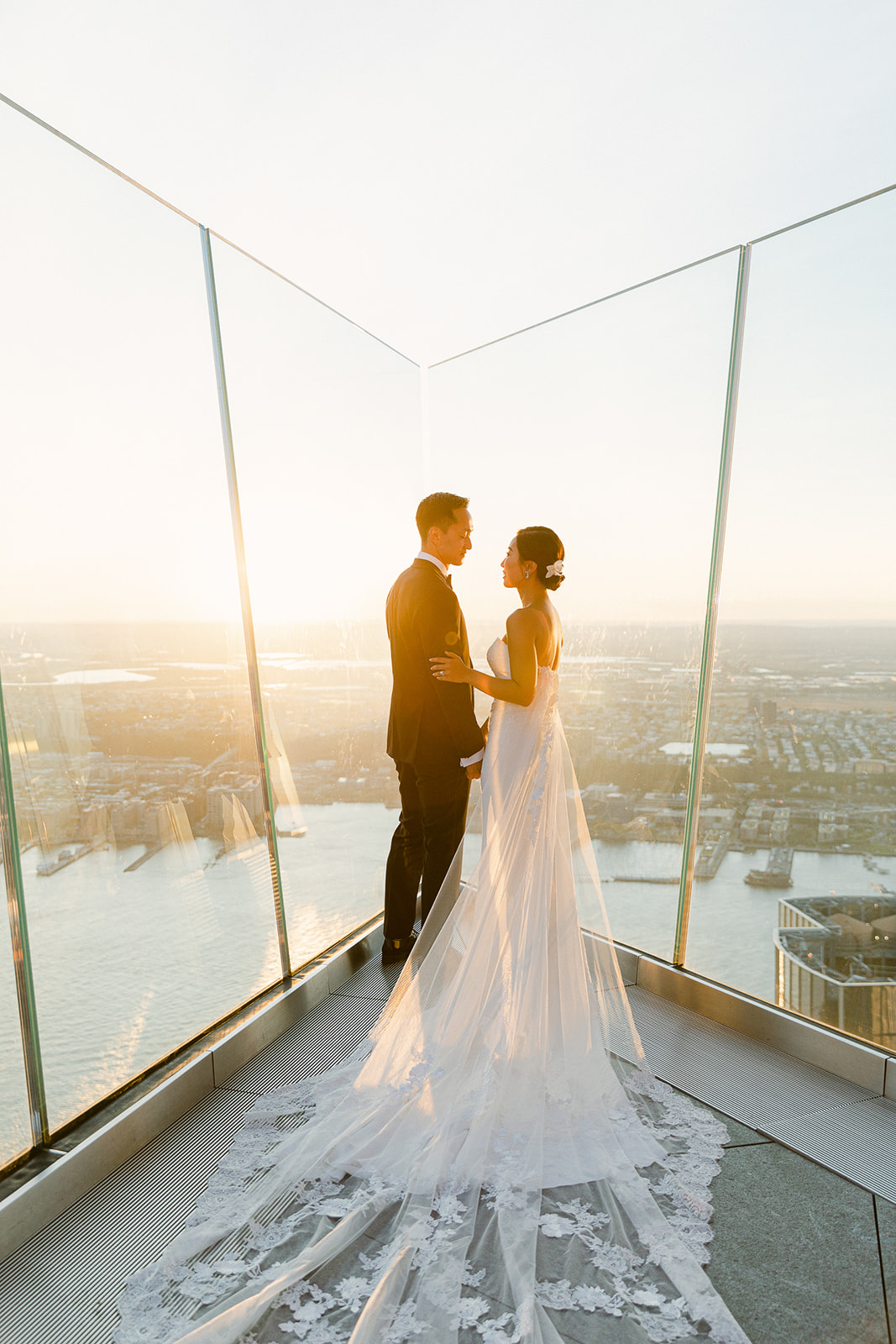 A bride and groom stand on a rooftop terrace at sunset, overlooking a scenic cityscape with a river. The bride wears a long, flowing white gown with lace detail. They are facing each other, embraced in a moment of togetherness.
