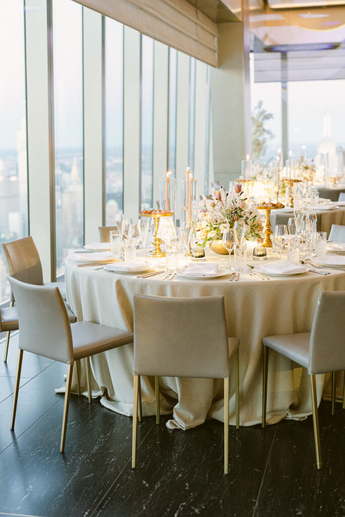 Elegant dining setup in a modern room with large windows. Round table with a white tablecloth, gray chairs, and golden candle holders. A floral centerpiece and glassware add sophistication. Cityscape visible in the background.