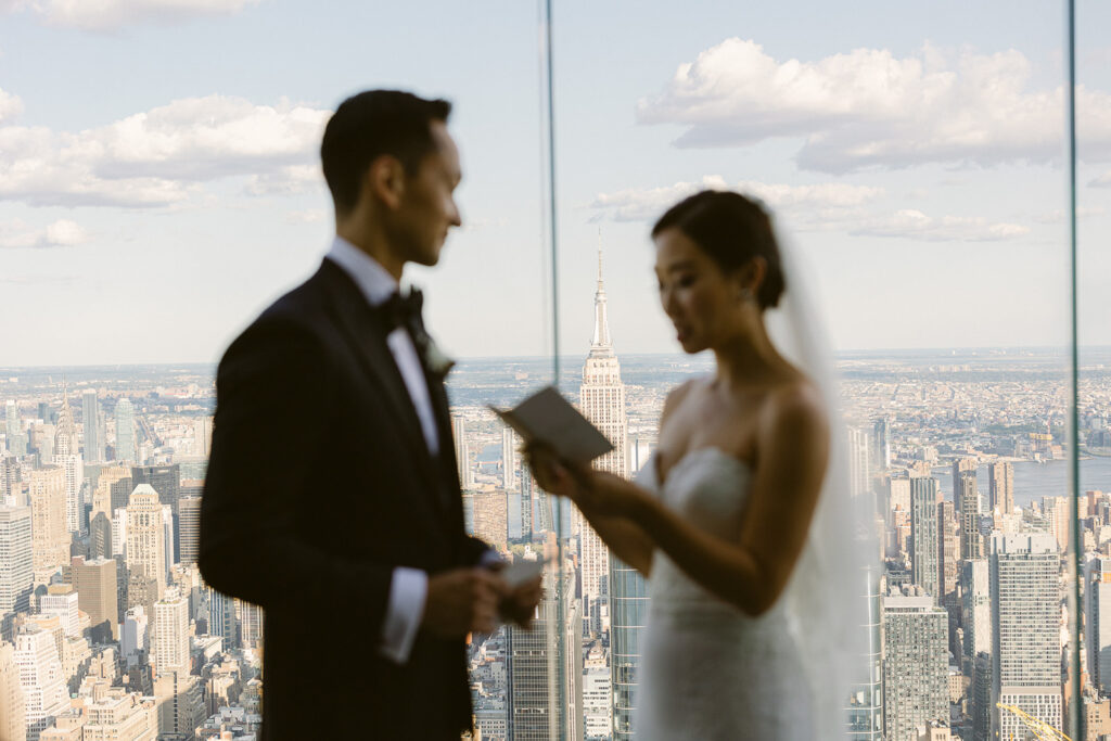 A couple stands inside a tall building with large windows, exchanging vows. The blurred cityscape of New York, including the Empire State Building, is visible in the background. The woman holds a small booklet, while the man listens attentively.