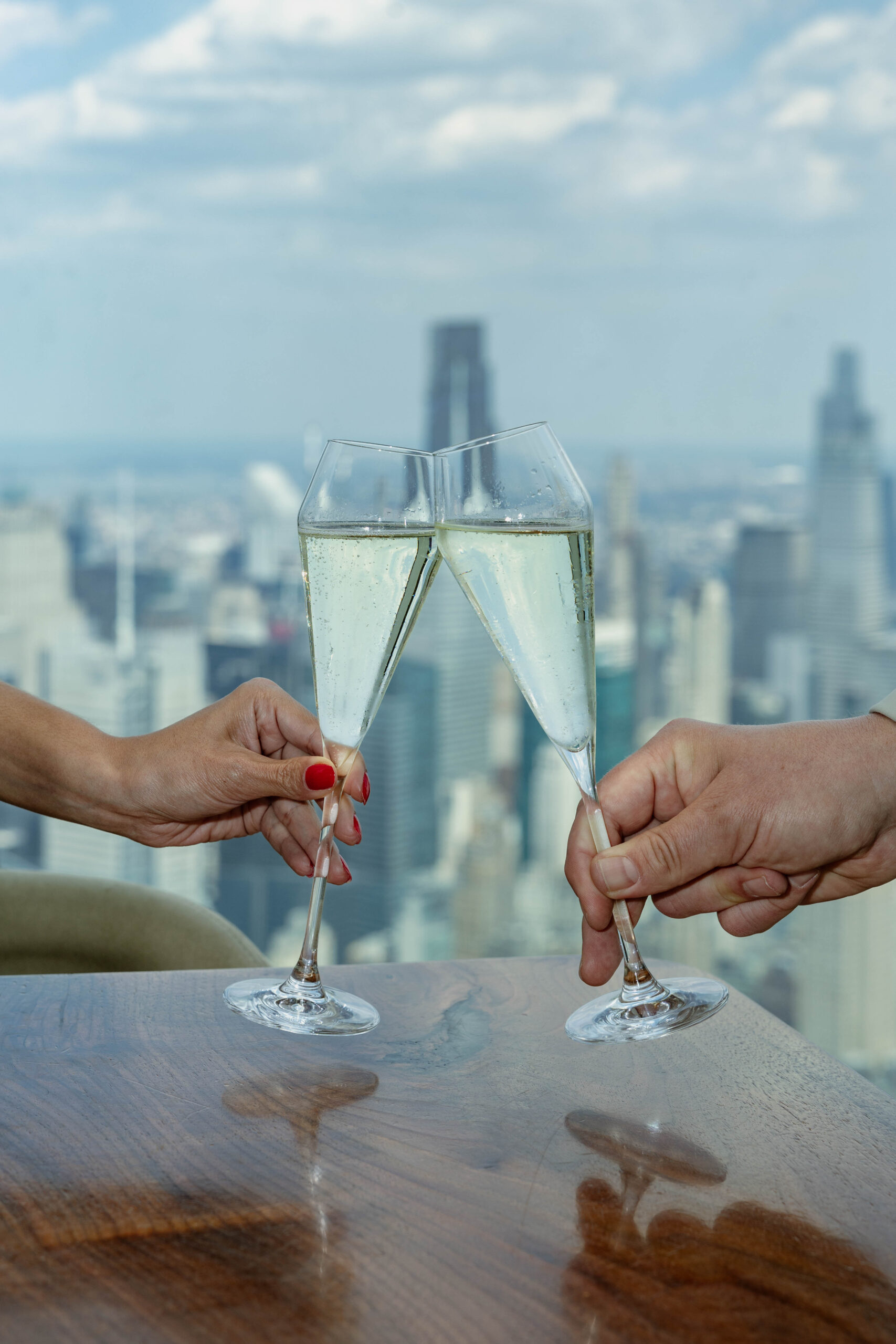 Two people clink champagne glasses over a wooden table, with a city skyline and tall buildings visible through a window in the background.