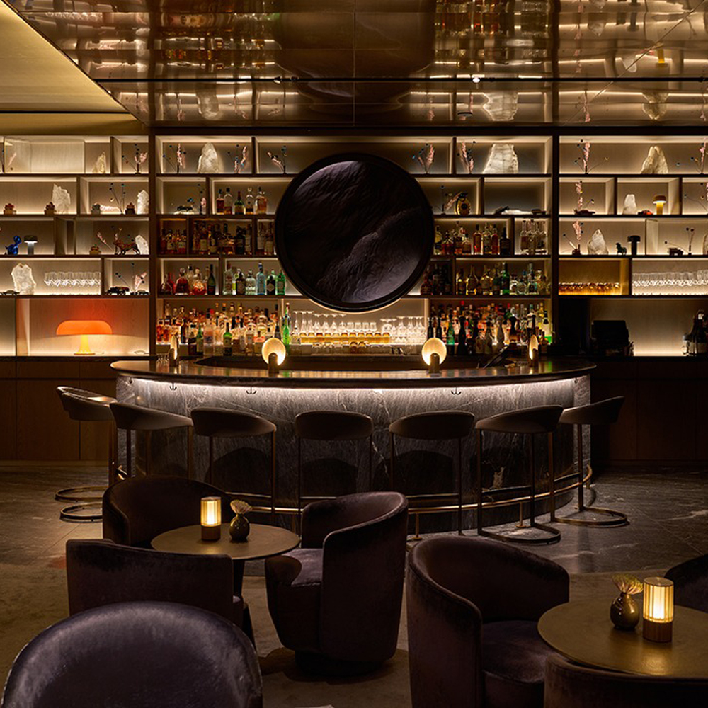 A stylish, modern bar with curved velvet seating, dim lighting, and a marble counter. Shelves behind the bar display bottles and glassware, illuminated by warm ambient lights and a large circular mirror as the centerpiece.